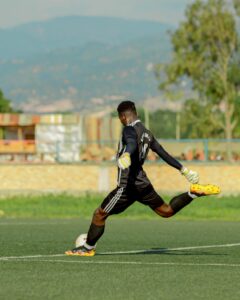 A football goalkeeper in action, kicking a ball on a sunny outdoor field.