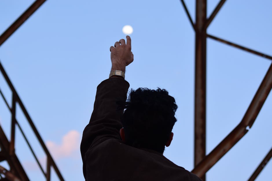 A man silhouetted against the morning sky reaches towards the moon with an outstretched hand.