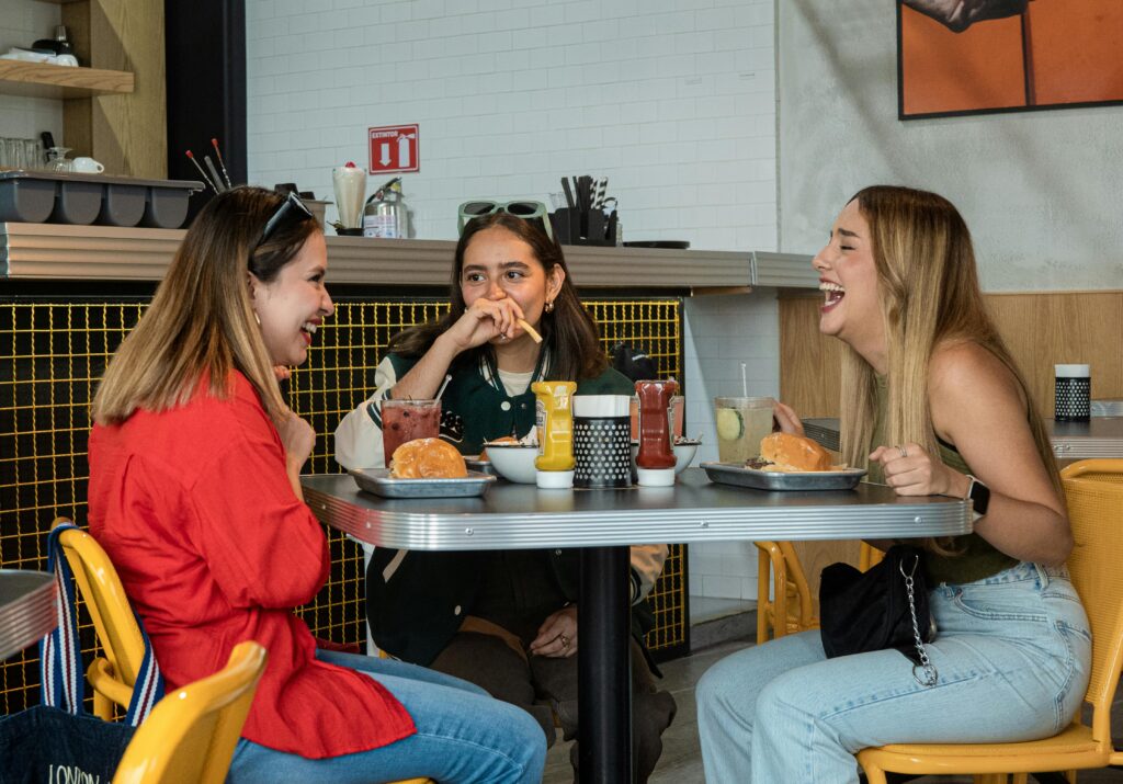 Three friends laughing and enjoying burgers in a vibrant city cafe.